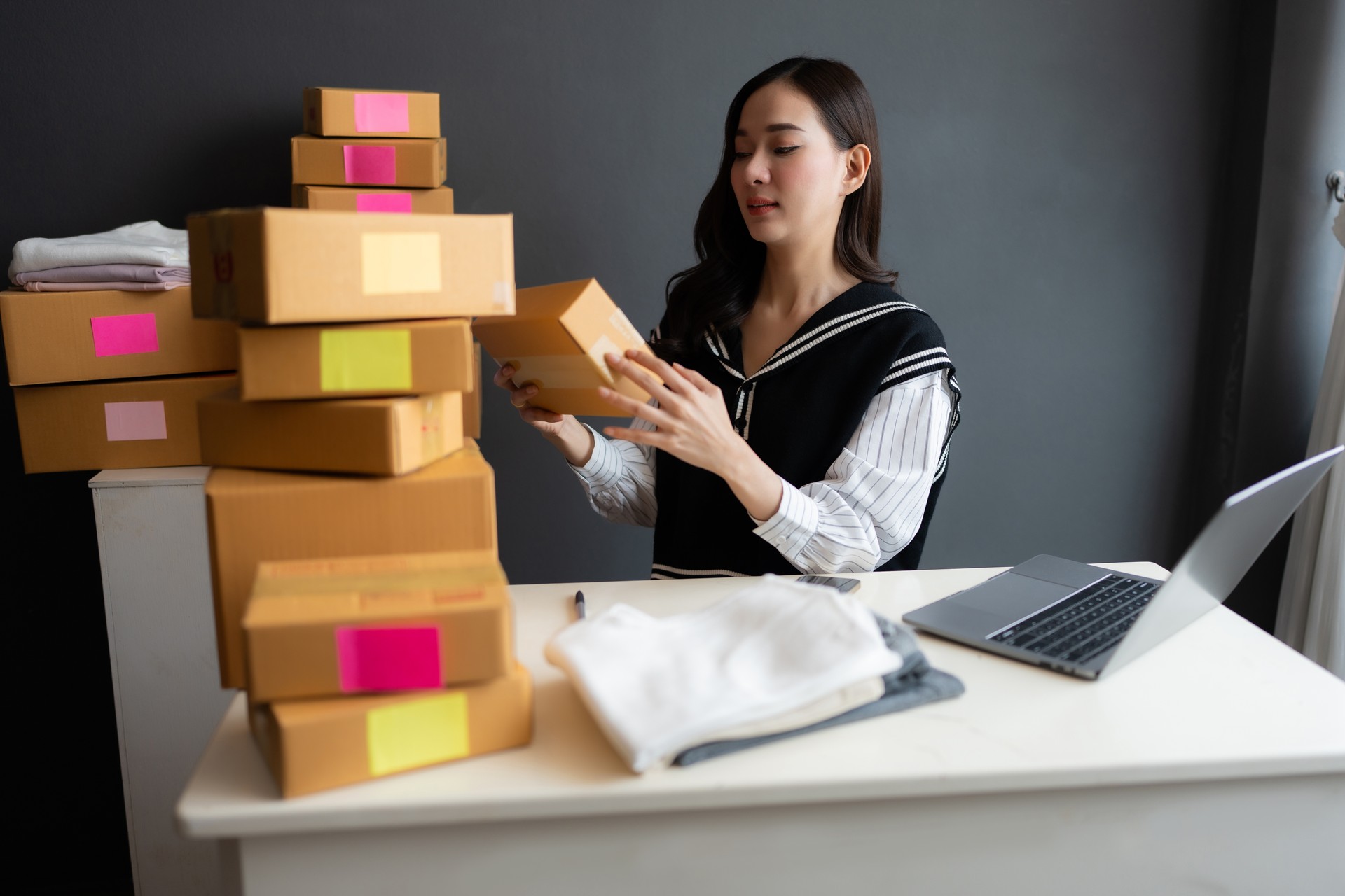 In the company's home office, there is an Asian female online seller who owns a startup business. Inspect and pack product orders into parcel boxes to prepare for delivery to customers.